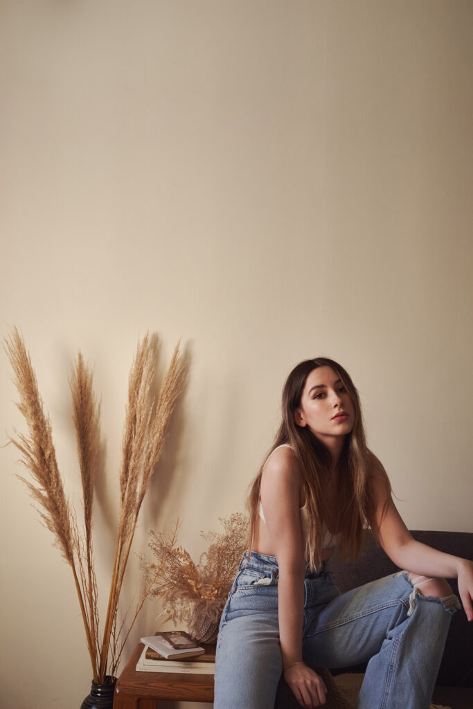 Gaby A serene portrait of a model seated on a couch with a backdrop of a beige wall decorated with tall dried grass and a wooden table holding books and a vase.